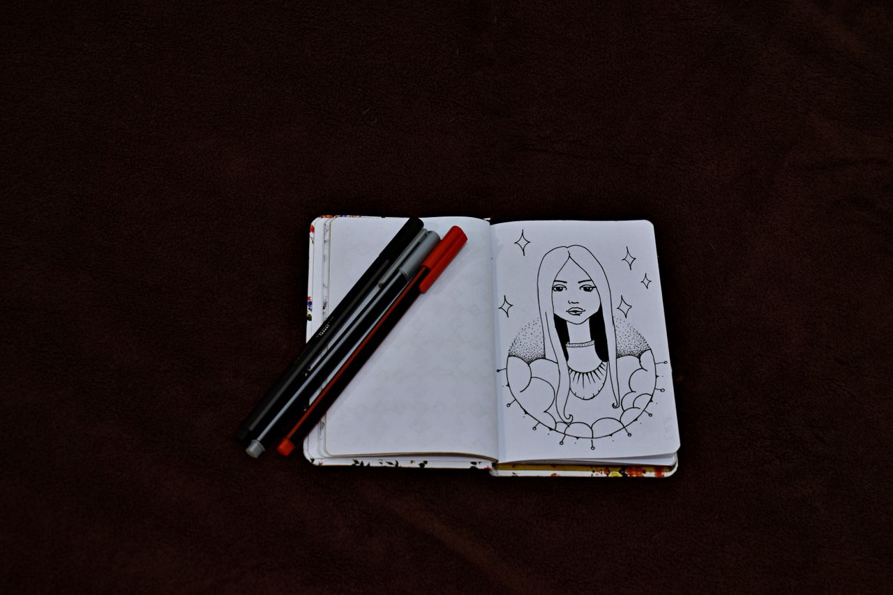 our-story-01 Open sketchbook with a detailed artwork and pens on a dark background, perfect for creative inspiration.